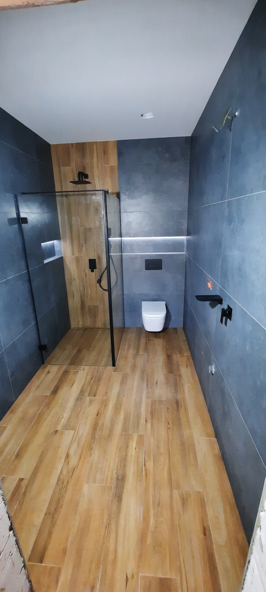 Nowoczesna łazienka z szarymi płytkami ściennymi, drewnopodobną podłogą, kabiną prysznicową walk-in z czarnymi profilami i wiszącą toaletą.