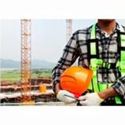 Pracownik budowlany w szarej koszuli w kratę i zielonych szelkach bezpieczeństwa trzyma pomarańczowy kask na tle budowy z dźwigami i oddalonym krajobrazem.