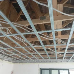 Konstrukcja metalowa podwieszanego sufitu z widocznymi przewodami elektrycznymi, zamontowana do drewnianej więźby dachowej w surowym wnętrzu.