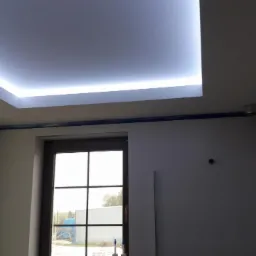 Pokój z podwieszanym sufitem z oświetleniem LED w kształcie prostokąta, listwy maskujące pomalowane na ciemny kolor, widoczne okno z widokiem na zewnątrz.