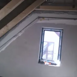 Poddasze w trakcie prac wykończeniowych: ściana po szpachlowaniu z osadzonym oknem, widoczne drewniane elementy konstrukcji dachu i przewody instalacyjne.