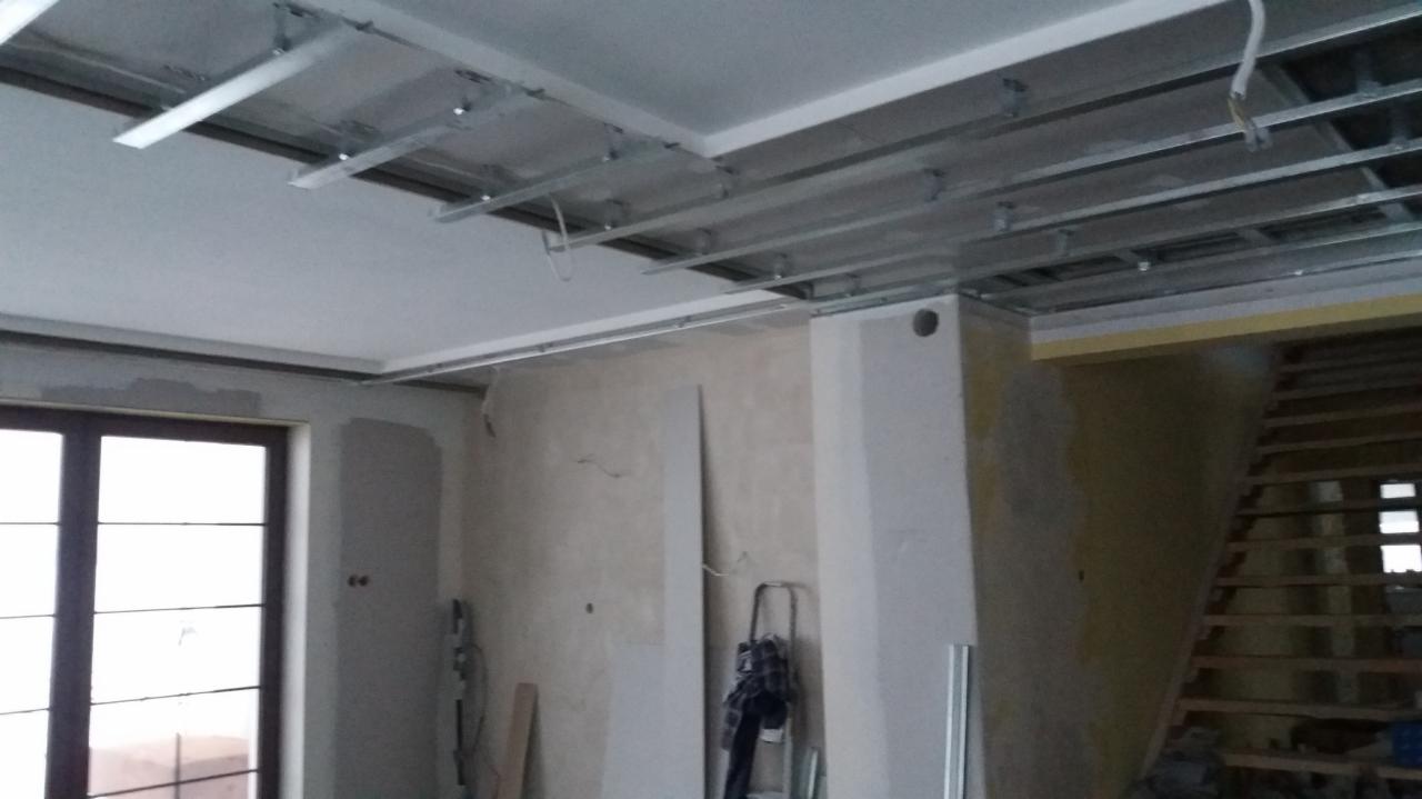 Montaż metalowej konstrukcji sufitu podwieszanego w trakcie remontu, widoczne profile, płyty gipsowe i drabina oparta o ścianę z widocznymi śladami po szpachlowaniu.