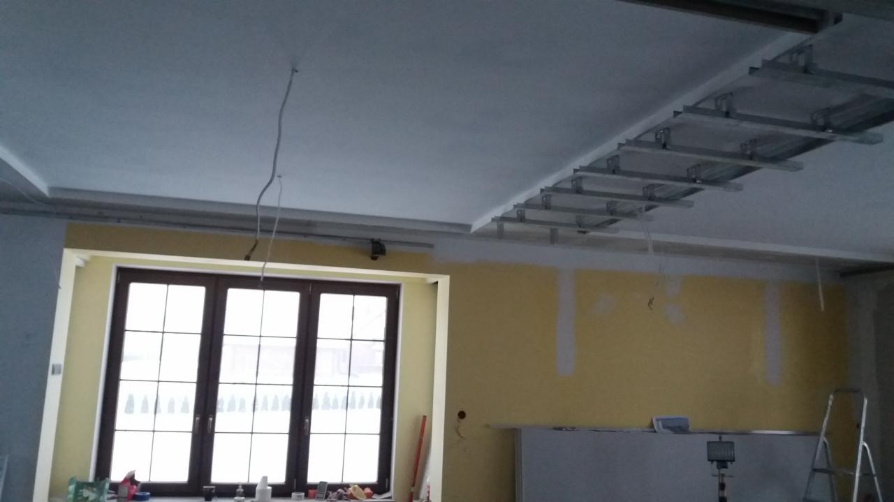 Wnętrze w trakcie remontu: widoczne profile metalowe do montażu sufitu podwieszanego, częściowo pomalowane ściany i okno z ciemną ramą.