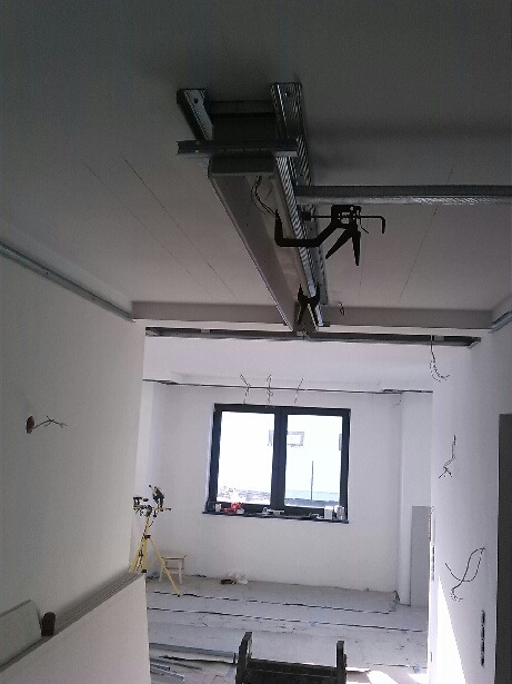 Montaż metalowego stelaża pod płytę kartonowo-gipsową na suficie w jasnym, pustym pomieszczeniu z oknem; widoczne narzędzia i przewody.