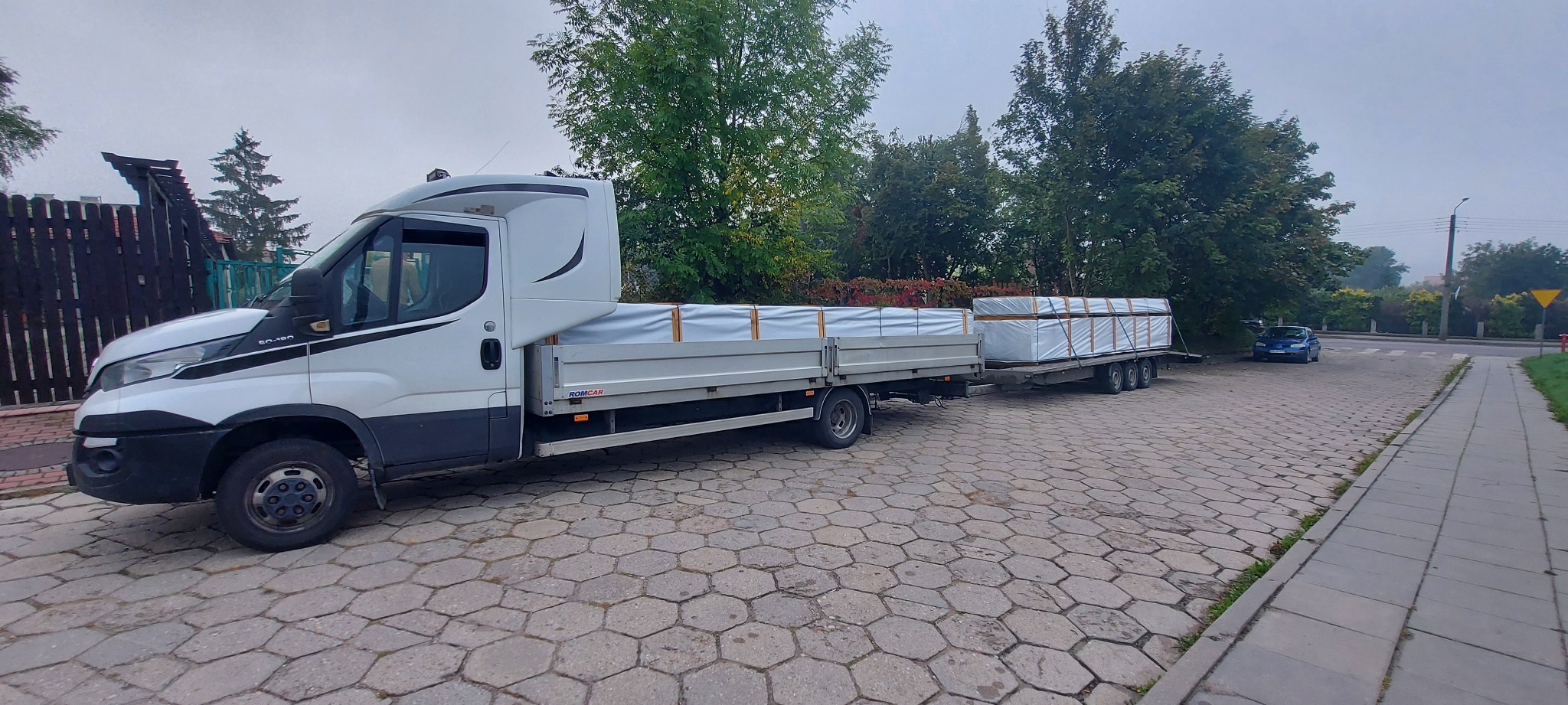 Biały bus z przyczepą przewozi ładunek zabezpieczony plandeką na brukowanym placu, widoczne drzewa i zaparkowany samochód osobowy w tle.