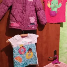 Różowa kurtka zimowa w kropki z kapturem i aplikacją Peppy Pig, obok niej koszulki dziecięce z różnymi nadrukami, wiszące na wieszakach na tle brązowych drzwi.