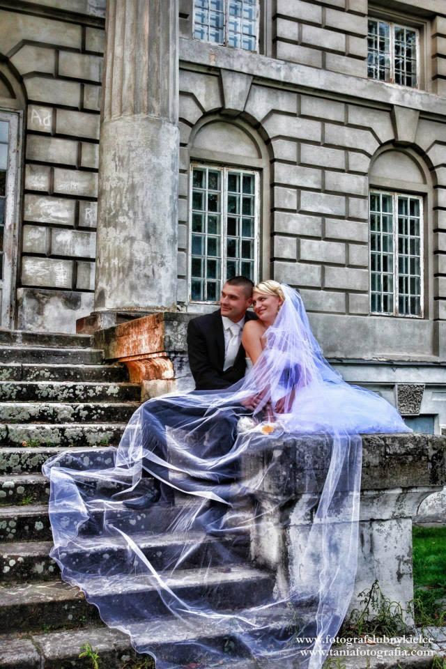 Para młoda siedzi na kamiennych schodach starego budynku, panna młoda w białej sukni z długim, przezroczystym welonem, pan młody w czarnym garniturze, oboje uśmiechnięci.