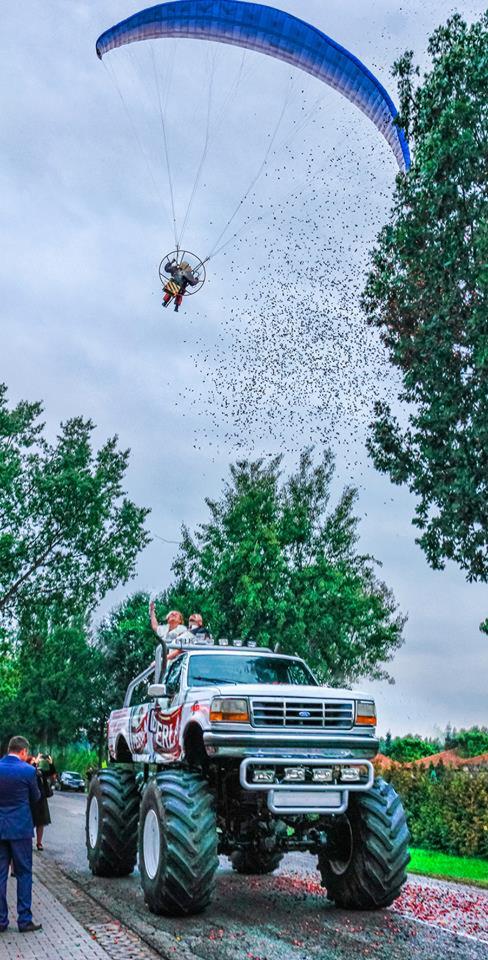 Monster truck z parą młodą, nad nimi paralotniarz zrzuca konfetti, w tle drzewa i goście weselni.