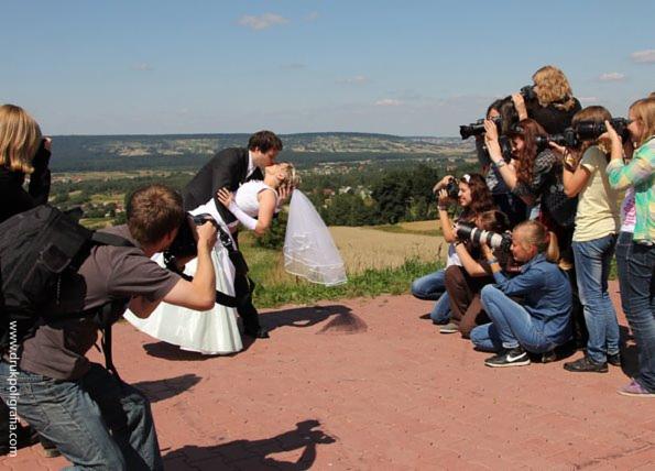 Para młoda w pocałunku, fotografowana przez grupę fotografów na wzgórzu z widokiem na krajobraz.