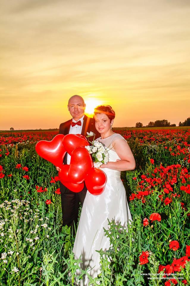 Para młoda w polu maków o zachodzie słońca, panna młoda trzyma bukiet i czerwone balony w kształcie serc, pan młody w smokingu z czerwoną muchą.