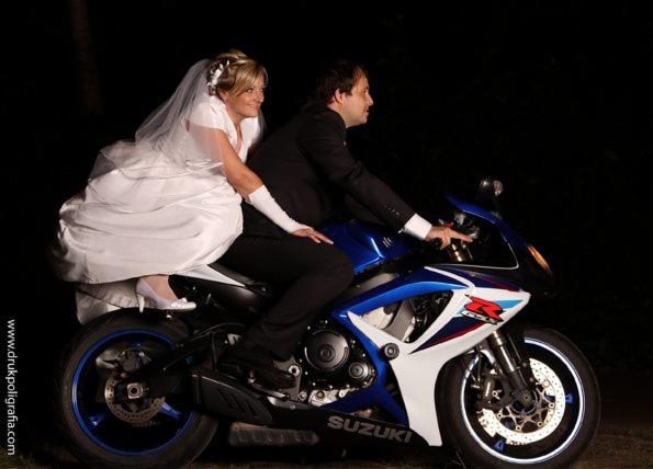 Panna młoda w białej sukni i welonie siedzi za panem młodym w garniturze na niebiesko-białym motocyklu Suzuki na ciemnym tle.