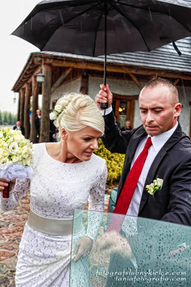 Panna młoda w koronkowej sukni i bukietem białych róż w deszczu, mężczyzna w garniturze z czerwoną kravatą trzyma nad nią czarny parasol, w tle budynek z drewnianymi kolumnami.