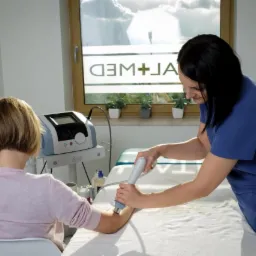 Pacjentka poddawana zabiegowi ultradźwiękami na przedramieniu przez terapeutkę w gabinecie z oknem i sprzętem medycznym.