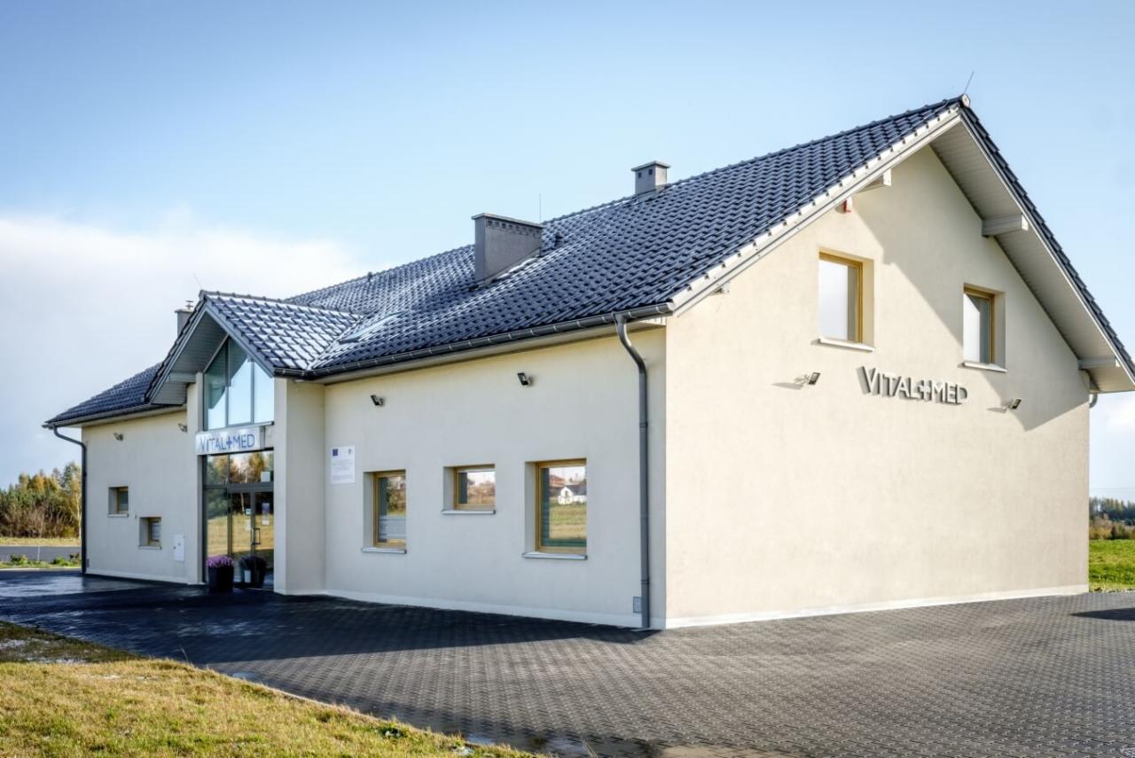 Budynek placówki medycznej Vitalmed z szarym dachem i jasną elewacją w słoneczny dzień. Widoczny trawnik i kostka brukowa przed wejściem.