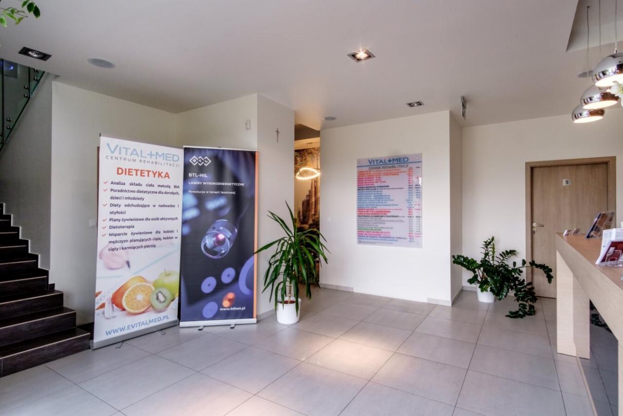 Jasne wnętrze centrum rehabilitacji z recepcją, schodami i roll-upami reklamowymi dietetyki i laseroterapii BTL-HIL.