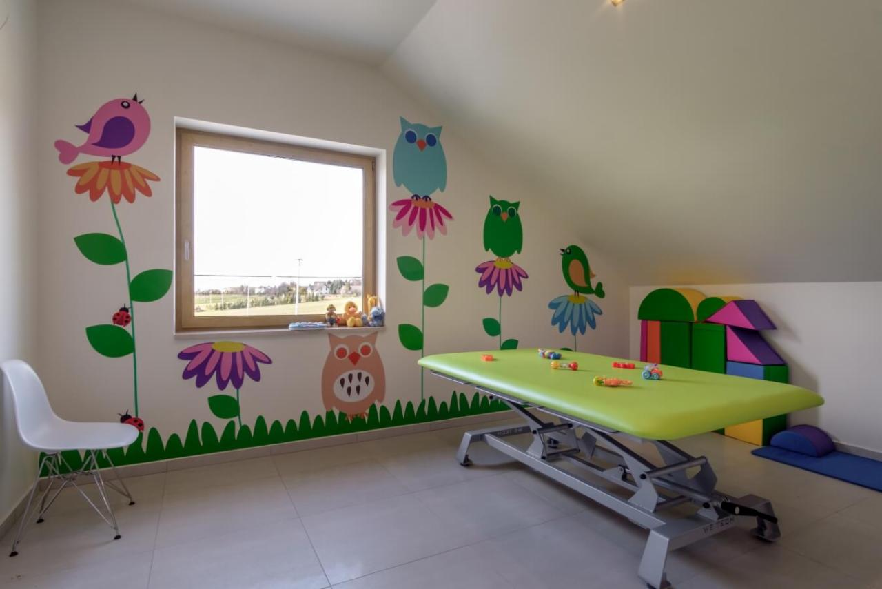 Jasny pokój z kozetką do ćwiczeń, ściany zdobione naklejkami z motywem kwiatów, ptaków i sów, zabawki na parapecie i stole.