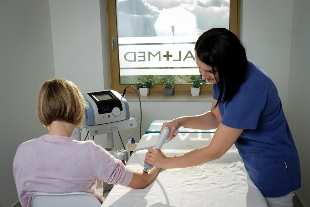 Pacjentka poddawana zabiegowi ultradźwiękami na przedramieniu przez terapeutkę w gabinecie z oknem i sprzętem medycznym.