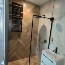 Nowoczesna łazienka z kabiną prysznicową typu walk-in z czarnymi profilami, drewnopodobnymi płytkami ułożonymi w jodełkę na ścianie i podłodze, oraz białą szafką pod umywalką z czarnymi uchwytami.