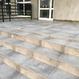 Wyłożone szarymi płytkami schody wejściowe do budynku z białą stolarką okienną i drzwiową, czarną balustradą oraz widocznymi śladami po pracach wykończeniowych na ścianie.