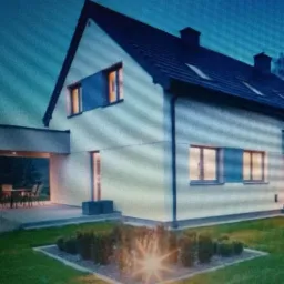 Nowoczesny dom jednorodzinny z białymi elewacjami, ciemnym dachem i oknami, z oświetlonym ogrodem i zadaszonym patio w tle, sfotografowany o zmierzchu.