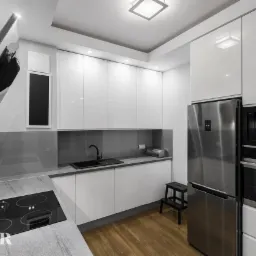 Nowoczesna kuchnia z białymi, lakierowanymi frontami, szarym blatem i drewnianą podłogą. Widoczny czarny zlew, płyta indukcyjna i stalowa lodówka.