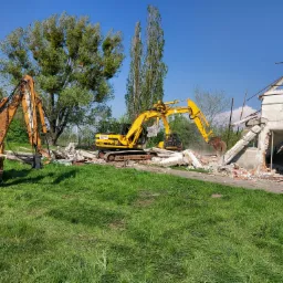 Dwa żółte i pomarańczowe koparki usuwają gruz z częściowo zburzonego budynku z czerwonej cegły na zielonym trawniku pod błękitnym niebem.
