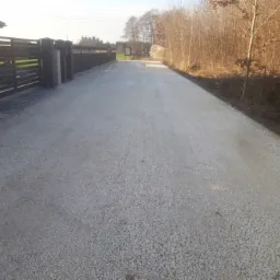 Utwardzona droga dojazdowa z jasnego kruszywa, zakończona montażem nowoczesnego, ciemnego ogrodzenia panelowego z betonowymi słupkami, prowadząca w kierunku lasu.