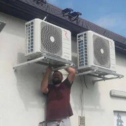 Uśmiechnięty monter instaluje dwie zewnętrzne jednostki klimatyzacyjne marki Rotenso na metalowych wspornikach przymocowanych do białej ściany budynku z brązowym dachem.