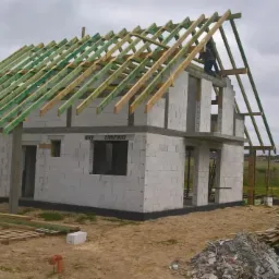 Budowa domu z białych bloczków: widoczna konstrukcja dachu z zielonych belek, robotnik na górze, tymczasowa toaleta WC Serwis na pierwszym planie.