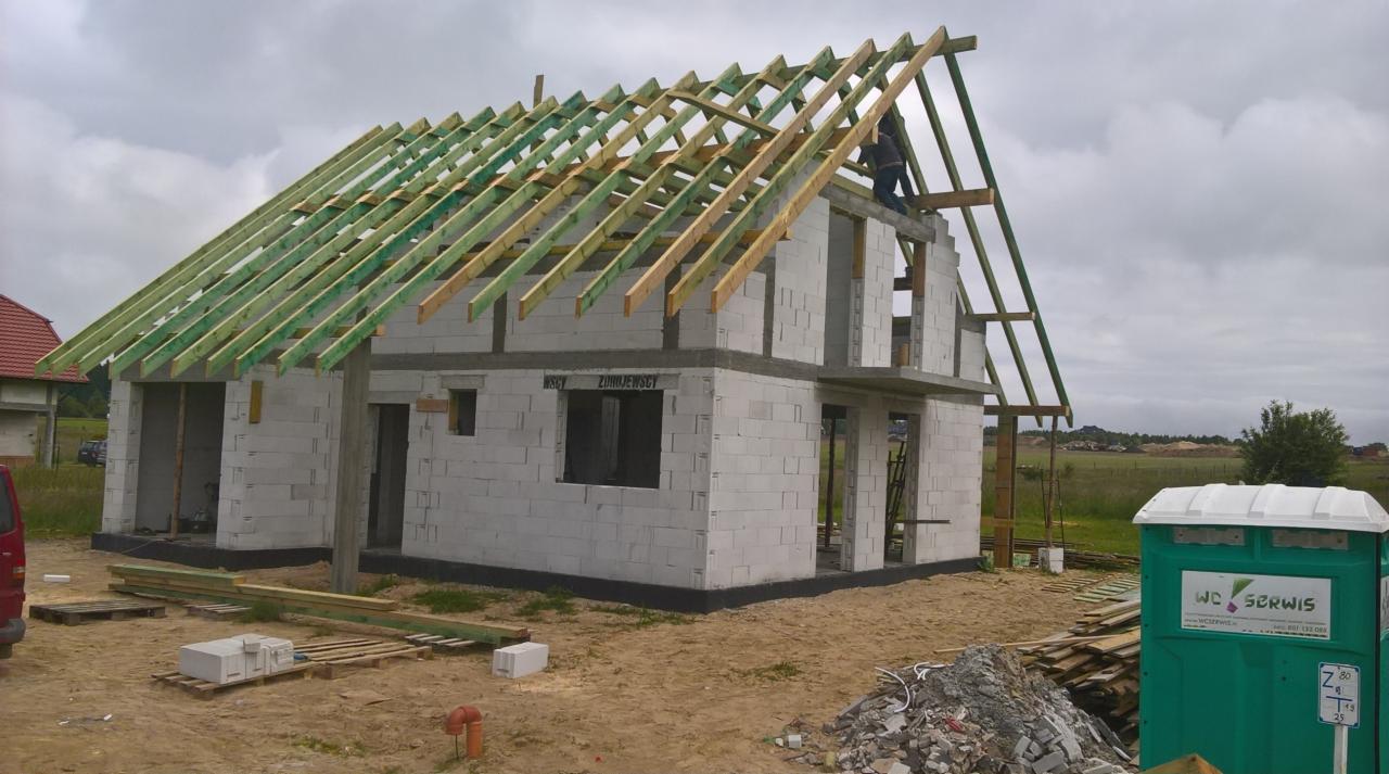 Budowa domu z białych bloczków: widoczna konstrukcja dachu z zielonych belek, robotnik na górze, tymczasowa toaleta WC Serwis na pierwszym planie.