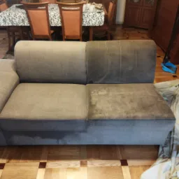 Szara kanapa dwuosobowa, z widoczną różnicą w czystości między połową wyczyszczoną a zabrudzoną, na parkiecie z ciemnymi wstawkami.