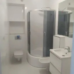 Jasna, minimalistyczna łazienka z kabiną prysznicową z szarymi szybami, wiszącą toaletą, umywalką z szafką i wnękami w ścianie wyłożonymi białymi płytkami.