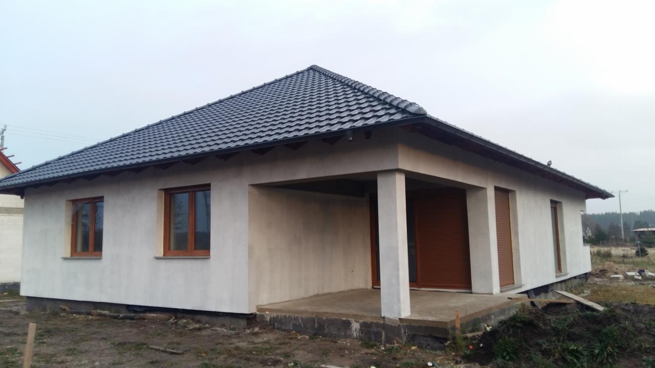 Niedawno wybudowany parterowy dom z ciemną dachówką, jasną elewacją w trakcie wykończenia i brązowymi oknami, widoczne nierówności terenu i elementy budowlane.