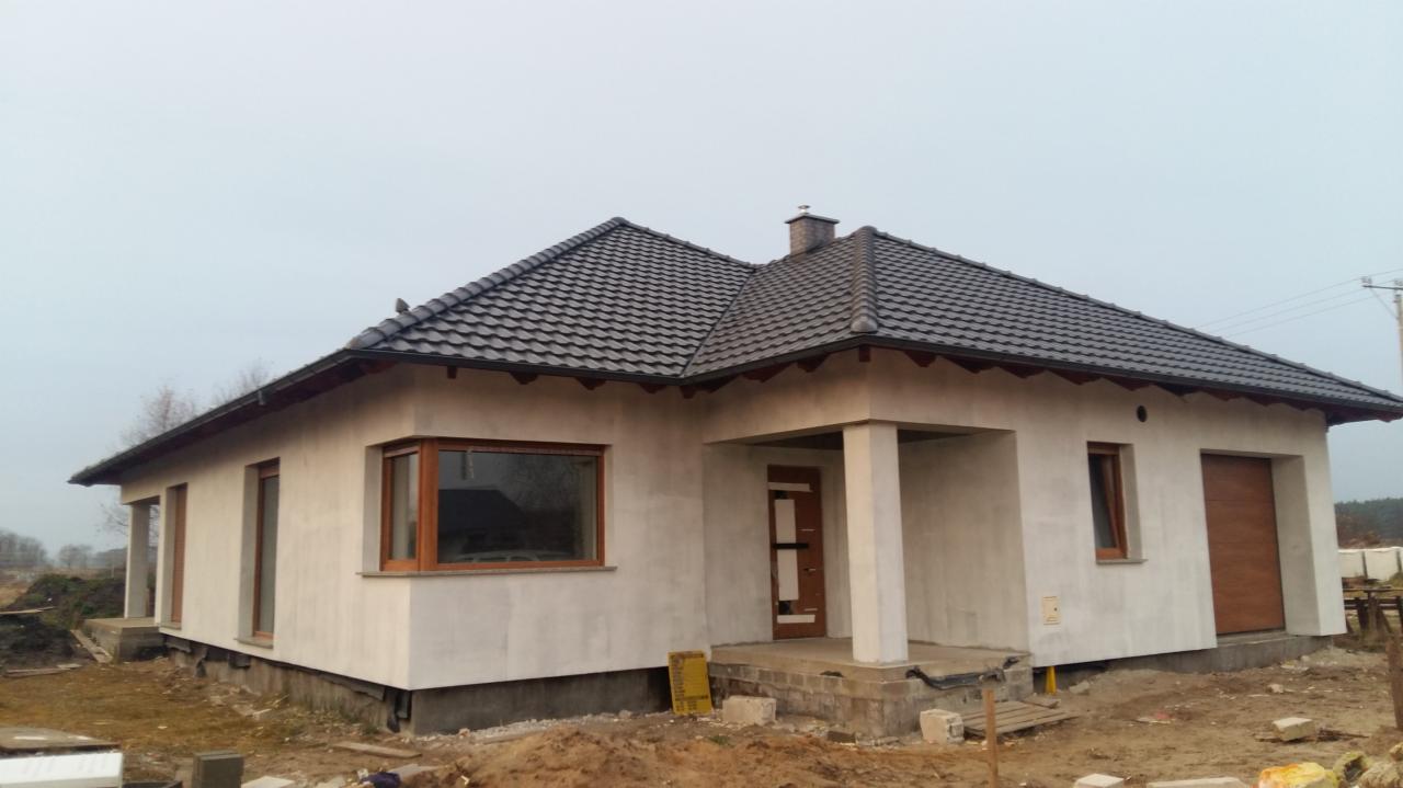 Niedokończony, parterowy dom jednorodzinny z szarą dachówką, drewnianą stolarką okienną i elewacją w trakcie wykończenia, widoczne niedoskonałości tynku i resztki materiałów budowlanych wokół budynku.