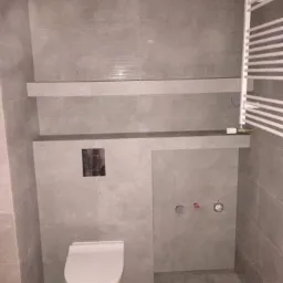 Nowoczesna, wykafelkowana łazienka w odcieniach szarości z podwieszaną toaletą, grzejnikiem drabinkowym i półkami w zabudowie. Widoczne wystające kable elektryczne na suficie.