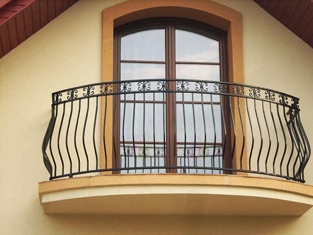 Balkon z czarną, metalową balustradą o falistym wzorze, umieszczony na beżowej elewacji domu z brązowym oknem w tle.