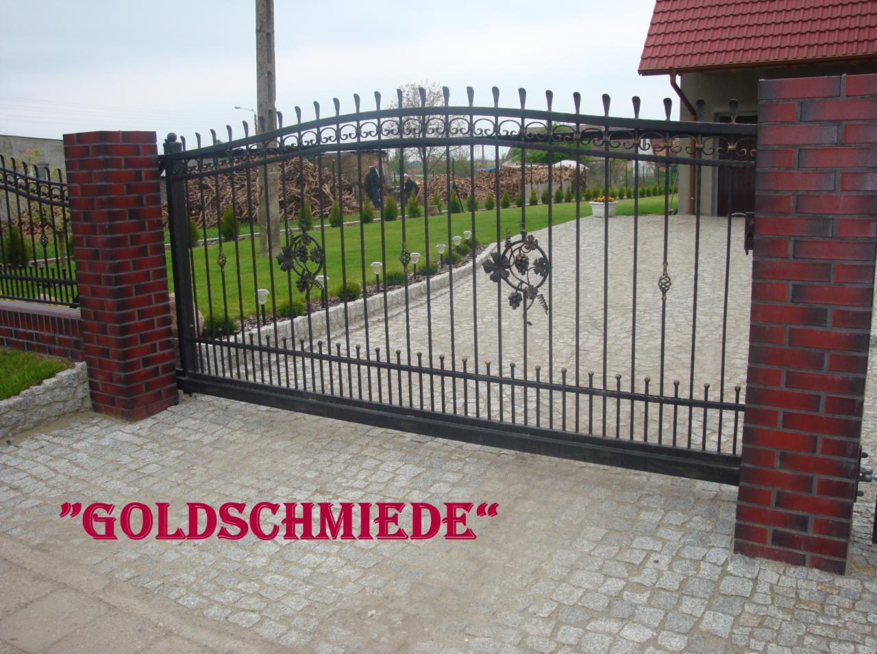 Czarna, metalowa brama wjazdowa z dekoracyjnymi elementami roślinnymi, osadzona między dwoma murowanymi słupkami z czerwonej cegły, na podjeździe z kostki brukowej.