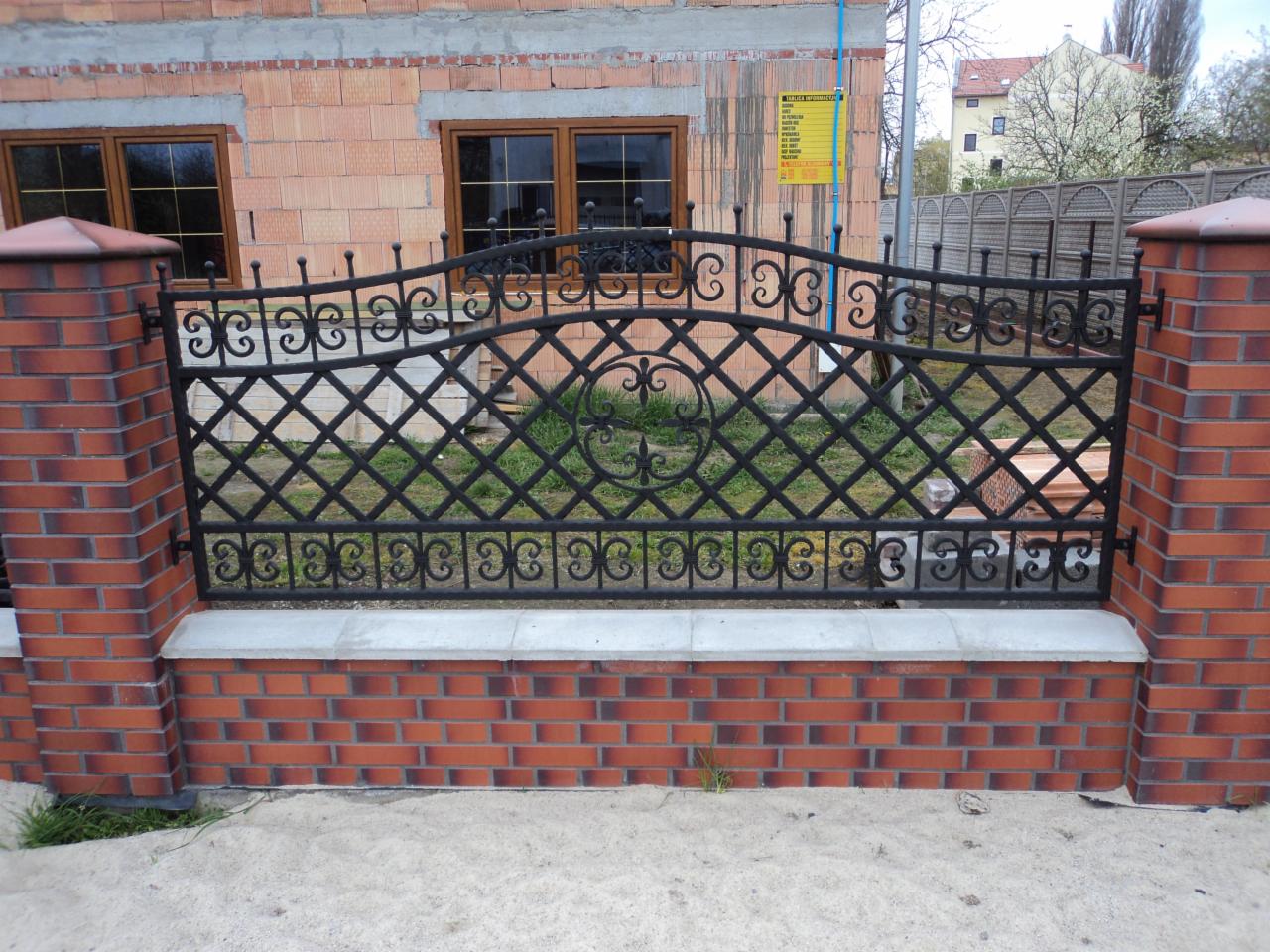 Kute, czarne ogrodzenie z ceglanymi słupkami, w tle budynek w trakcie budowy z oknami z brązowymi ramami.