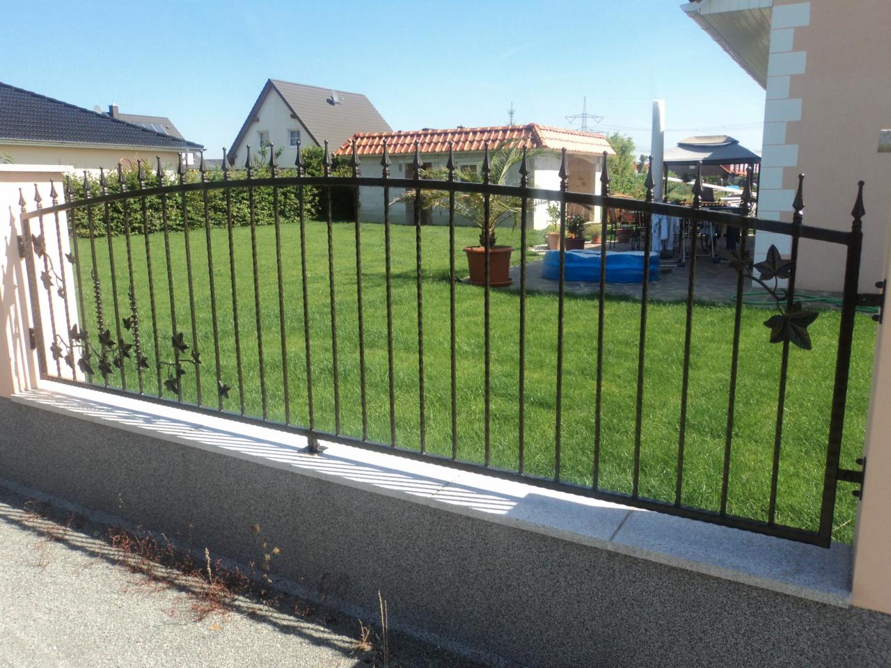 Kute, metalowe ogrodzenie z motywem liści na niskiej podmurówce oddziela zadbany trawnik z domkiem ogrodowym w tle.