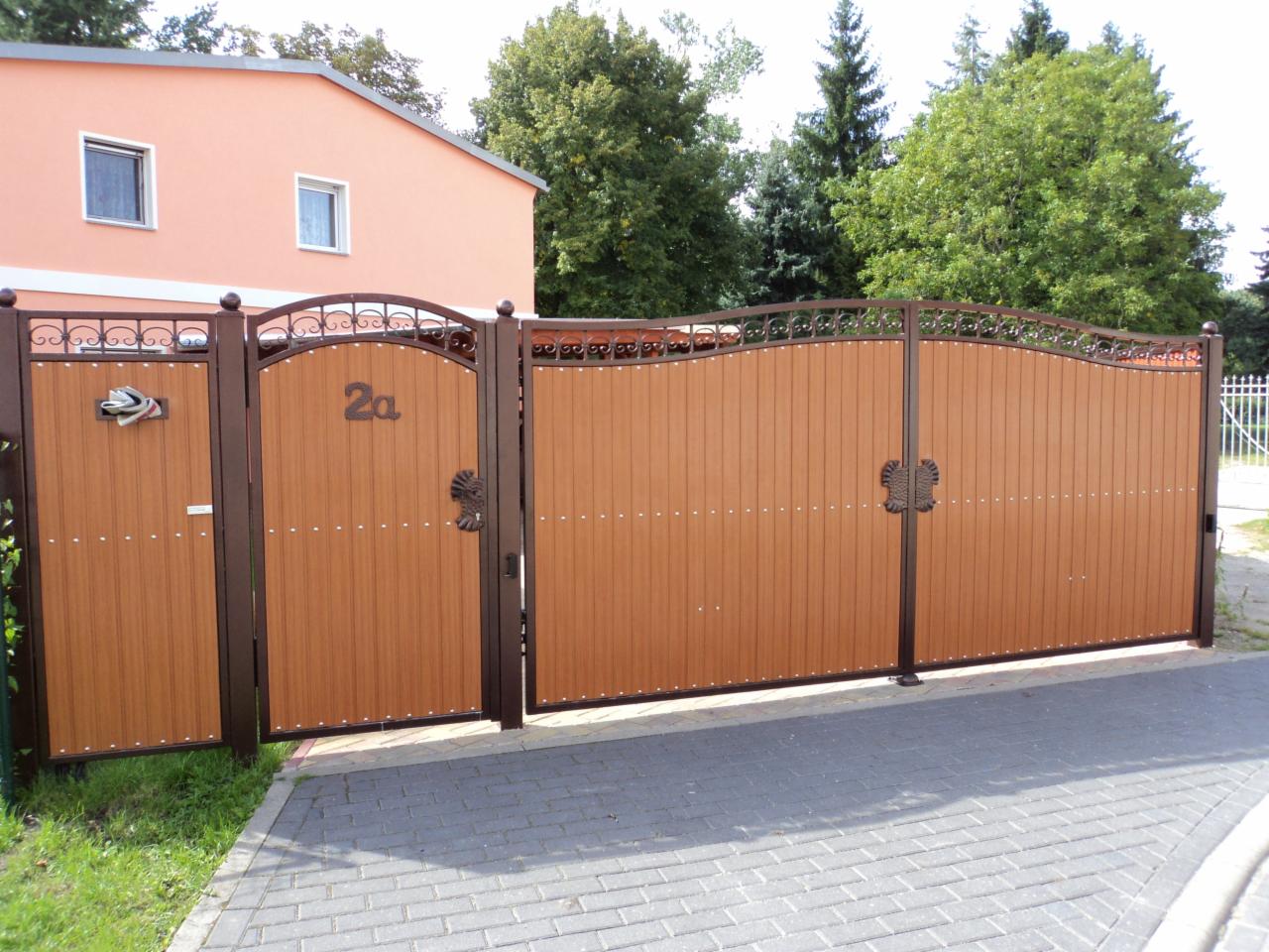 Brązowa brama wjazdowa z furtką, zdobiona metalowymi elementami i numerem domu '2a', na tle jasnego budynku i zieleni.