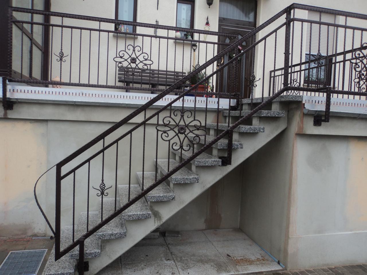 Zewnętrzne schody z szarego granitu prowadzące do drzwi wejściowych, zabezpieczone metalową balustradą z dekoracyjnymi elementami w kolorze ciemnego brązu.