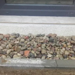 Kamienny drenaż u podstawy ściany budynku z oknem, widoczny fragment elewacji o ziarnistej fakturze i szarej podmurówki.