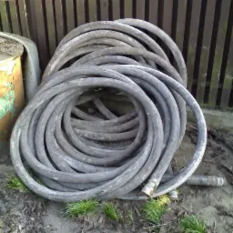 Zwinięty wąż do betonu leżący na ziemi, obok blaszany pojemnik i drewniane ogrodzenie.