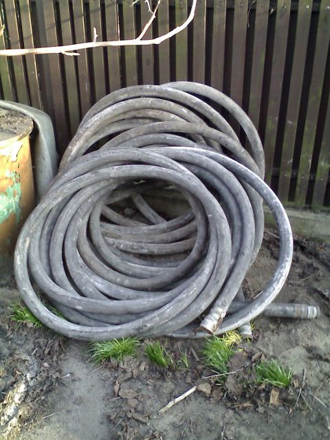 Zwinięty wąż do betonu leżący na ziemi, obok blaszany pojemnik i drewniane ogrodzenie.