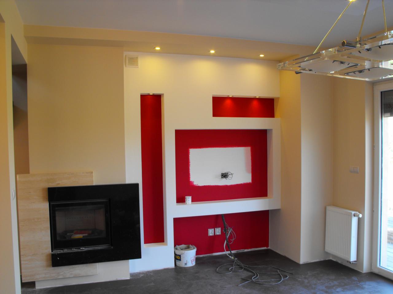Wnętrze pokoju z kominkiem i fragmentem ściany z wnękami pomalowanymi na czerwono, widoczne niedokończone malowanie i kable wystające ze ściany.