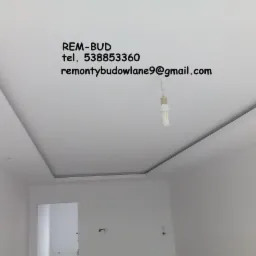 Wykończony sufit podwieszany z wnęką oświetleniową, z widocznym adresem e-mail i numerem telefonu firmy REM-BUD na g&oacute;rze kadru, w tle surowa żar&oacute;wka wisząca na kablu.
