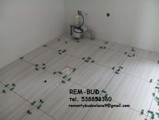 Białe płytki podłogowe w trakcie układania, z zielonymi krzyżykami dystansowymi, widoczny odpływ liniowy, w tle białe ściany i wiadro z narzędziami.