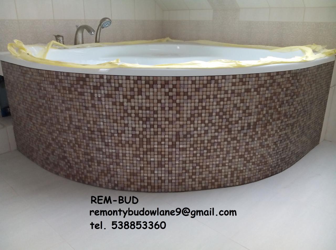 Owalna wanna obudowana mozaiką w odcieniach brązu, zabezpieczona folią malarską, z widoczną baterią i słuchawką prysznicową na tle jasnych płytek.