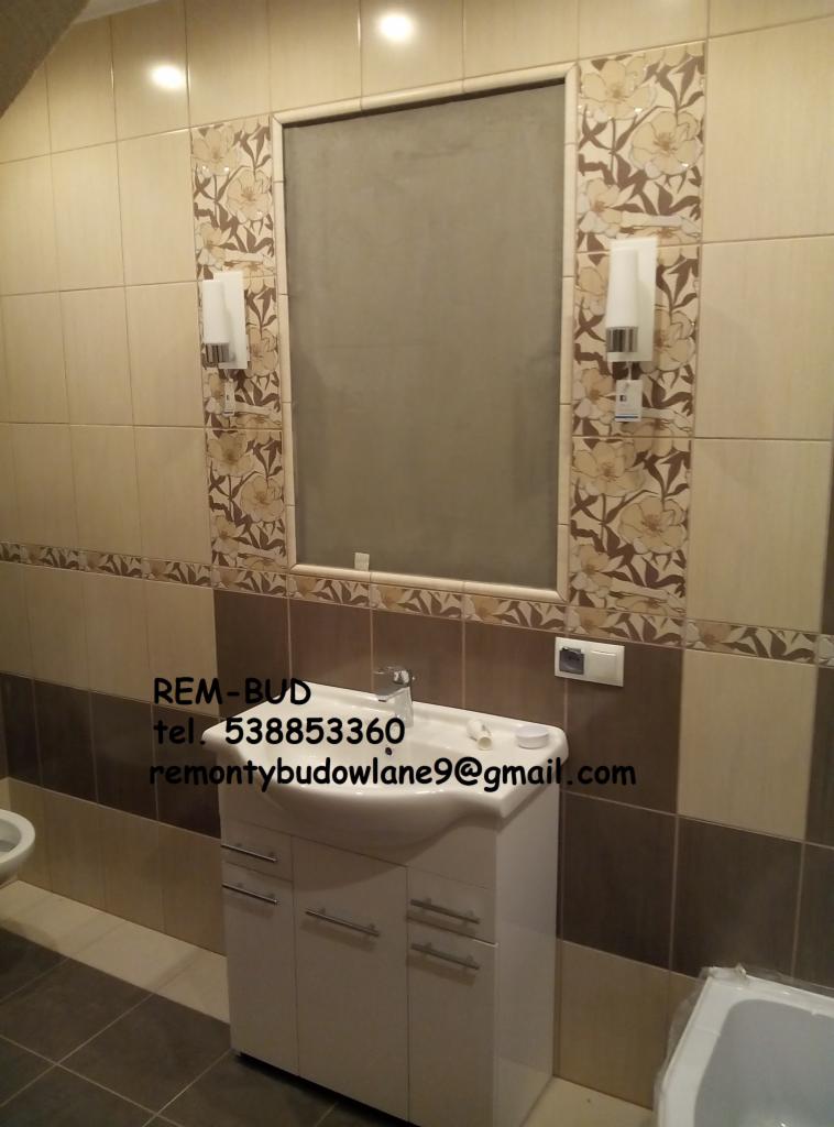 Ukończona łazienka z beżowo-brązowymi płytkami, umywalką z szafką i lustrem z dekoracyjnym obramowaniem, oświetlona dwoma kinkietami. Na umywalce biały pojemnik i rurka.