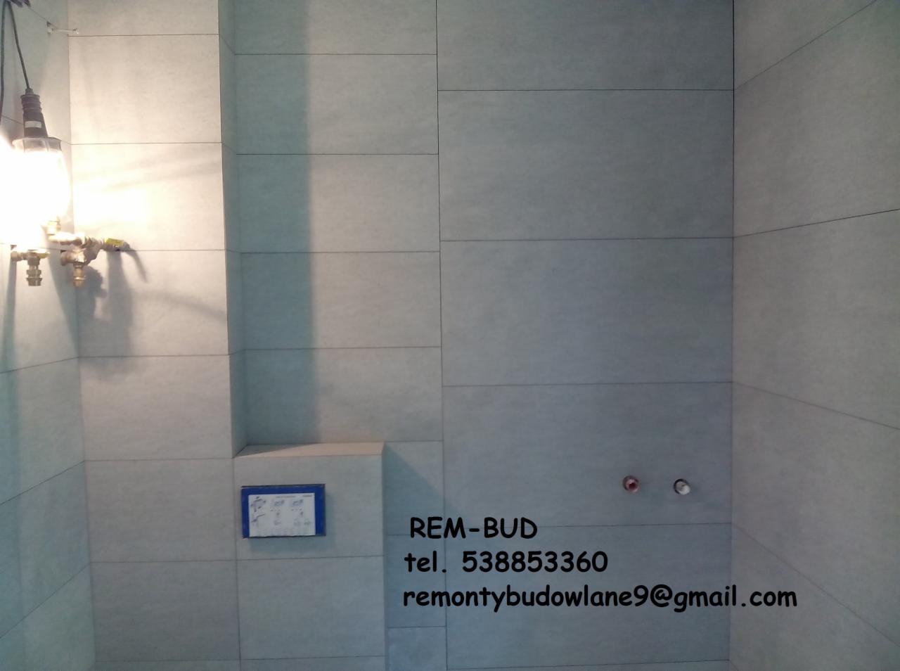 Świeżo wyłożona, szara, prostokątna glazura ścienna w łazience, z widocznymi przyłączami hydraulicznymi, wiszącą lampą roboczą i danymi kontaktowymi firmy REM-BUD.